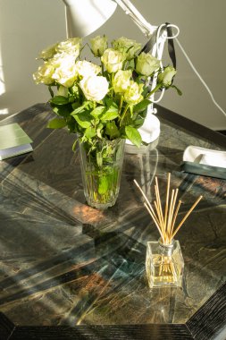 Taze beyaz güllerden oluşan bir vazo ve güneş ışığıyla aydınlatılmış, koyu ahşap bir masaya yerleştirilmiş bir cam kamış ayırıcı. Gölgeler rahat ve zarif bir atmosfer yaratır..