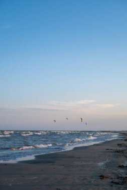 Okyanus dalgalarının kıyıya vurduğu kumlu bir sahil. Uçurtmacılar gün batımında gökyüzünde süzülürken. Pastel gökyüzü suyu yansıtır, dinamik ama dinamik bir kıyı manzarası yaratır..