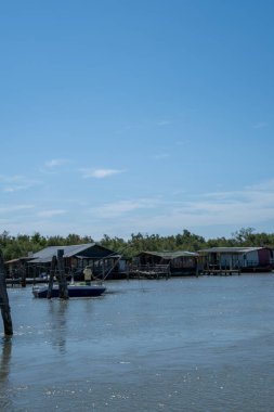 Sarı gömlekli bir adam küçük bir teknede nehir kıyısındaki ahşap yüzen evlerin yanında balık tutuyor. Sakin su, berrak mavi gökyüzünü yansıtıyor ve sakin bir manzara yaratıyor..