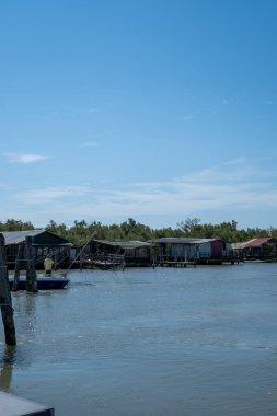 Sarı gömlekli bir adam küçük bir teknede nehir kıyısındaki ahşap yüzen evlerin yanında balık tutuyor. Sakin su, berrak mavi gökyüzünü yansıtıyor ve sakin bir manzara yaratıyor..