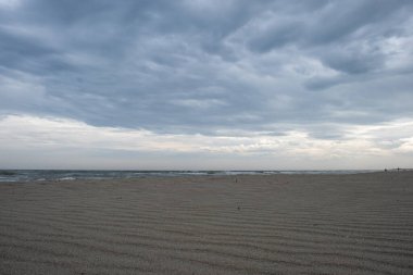 Bulutlu bir gökyüzünün altında kumlu bir kumsalın alçak açılı görüntüsü. Ön planda bir deniz kabuğu uzanıyor, doğal bir element ekliyor..