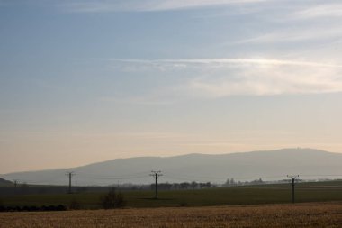 Elektrik hatları sisli tarlalar ve yumuşak gündoğumu ışığı altında uzak tepeler boyunca uzanan kırsal alan..