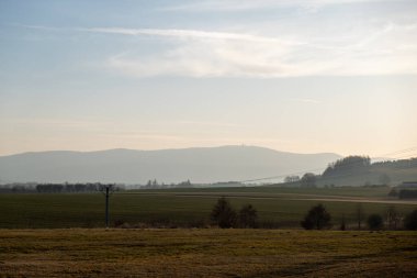 Geniş kırsal manzara, sisli tepeler, açık alanlar ve yumuşak bir sabah gökyüzünün altında elektrik direkleriyle..