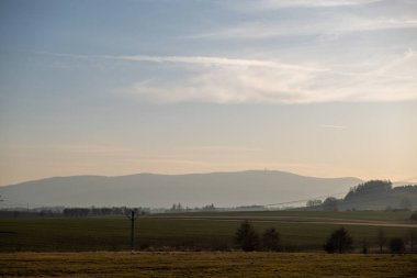 Geniş kırsal manzara, sisli tepeler, açık alanlar ve yumuşak bir sabah gökyüzünün altında elektrik direkleriyle..