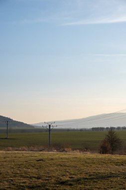 Geniş kırsal manzara, sisli tepeler, açık alanlar ve yumuşak bir sabah gökyüzünün altında elektrik direkleriyle..