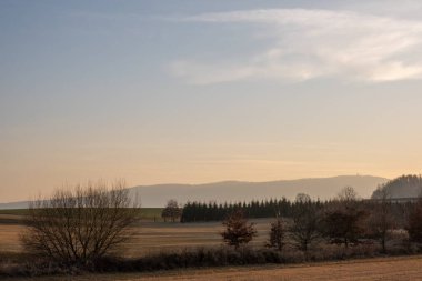 Kuru tarlalar, dağınık ağaçlar ve sıcak günbatımı ışığında uzak tepelerle sakin bir kırsal alan.