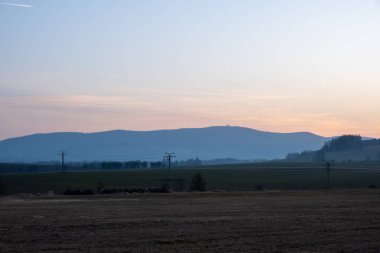 Kırsal alanda alacakaranlıkta sisle kaplı tepeler ve elektrik direklerinin siluetleri açık alandan görülüyor.