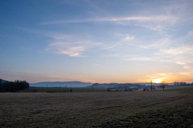 Açık kırsal arazinin üstündeki renkli günbatımı gökyüzü ve ilkbaharın başlarında görülen elektrik direkleriyle uzak tepeler.