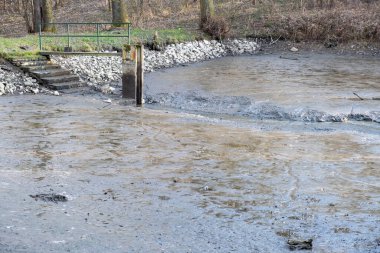 Baharın başında ormanlık alanda küçük beton baraj ve merdivenleri olan boş çamurlu bir gölet.