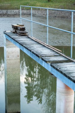 Yansıtıcı su yüzeyinin üzerinde metal parmaklıklar ve ahşap tahtalar olan bir yaya köprüsüne yakın plan, istiflenmiş tahtaları gösterir..