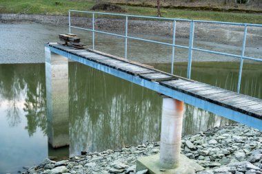 Yansıtıcı su yüzeyinin üzerinde metal parmaklıklar ve ahşap tahtalar olan bir yaya köprüsüne yakın plan, istiflenmiş tahtaları gösterir..