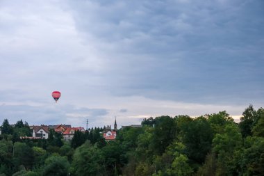 Avrupa 'nın bulutlu bir köyünün üzerinde uçan renkli sıcak hava balonu ve arka planda ağaçlar, geniş manzara manzarası.