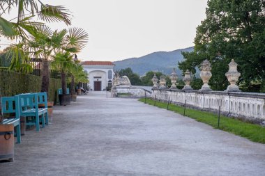 Tahta fıçılarda palmiye ağaçları olan taş teras, turkuaz banklar, ve barok balustrade, arka planda dağ manzarası.
