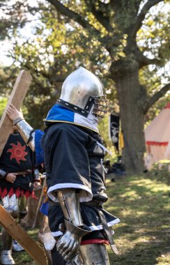 Ortaçağ şövalyesi kostümü giymiş bir adam. Adam savaştan sonra üzerinde demir zırh, büyük parlak bir kask, mavi gömlek ve demir eldivenlerle yürüyor.
