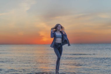 Bir su kütlesinin yanında duran biri. Yüksek kaliteli bir fotoğraf, ince bir kız gün doğumunda denizin sahilinde duruyor. Dar kotlar giyiyor ve boyunu aşmıyor. 