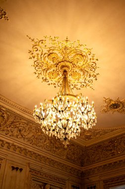 A huge crystal flake hangs on the stucco ceiling. The big chandelier hangs down. the chandelier shines, made of crystal. around the chandelier, on the ceiling stucco in baroque style clipart