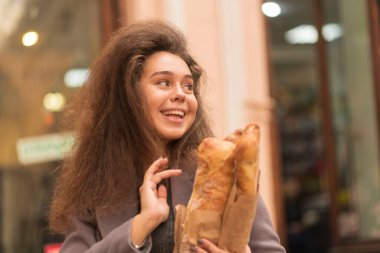 Caddedeki güzel, genç bir kadın kafenin baget ekmeği var. Kızların yüzüne yakından bakınca, başka yere bakıyor. Tatlı ve açık bir gülümsemesi var. Kabarık, kalın saçları var. Gri bir ceket giymiş.