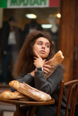 Genç bir kız bir kafede bir masada oturuyor ve bir ekmeği var. Kız başka tarafa bakıyor. O, arkadaşını bekliyor. Üç parça baget ekmeği var. Öğrencinin dalgalı kabarık saçları var..
