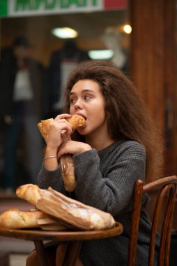 Genç bir kız bir kafede bir masada oturuyor ve bir baget yiyor. Bir baget yiyor ve onu ısırıyor. Onun önündeki masada üç baget ekmeği var. Öğrencinin dalgalı kabarık saçları var..