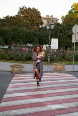 Kızın biri elinde bir baget ile yaya geçidinde karşıdan karşıya geçiyor. Elinde bir baget ile karşıdan karşıya geçen Coata 'lı genç bir öğrenci. Saçları kabarık.