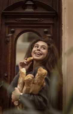 Kızın baget ekmeği var. Ortalama çekim, kameraya gülümsüyor. Ellerinde üç baget ve bir tane var.