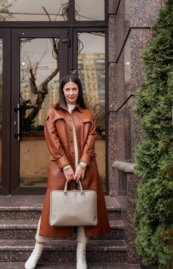 40 yaşın üzerinde zarif bir kadın laptop çantasıyla binadan çıkıyor. O kahverengi bir ceket giyiyor, dizüstü bilgisayar çantası tutuyor. O, beyaz ayakkabılar içinde zarif. O bir ofiste çalışan ince, bakımlı bir kadın..