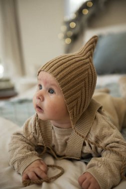 Şapka takan bir bebek. Küçük bir çocuk yatakta sürünüyor. Annesi onun arkasında oturuyor. Küçük çocuk bej bir şapka örüyor. Bebek çok hareketli ve sevimli, yatakta sürünüyor..
