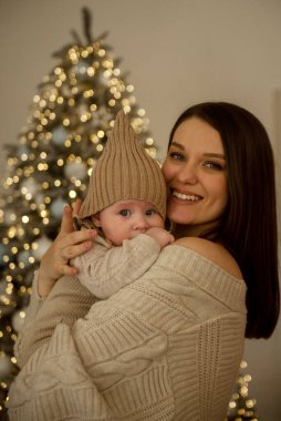 Bebek tutan bir kadın. Kız çocuğunu kollarında tutuyor. Çocuk bej bir el örgüsü şapka giyiyor. Anne ve çocuk mutlu, gülümsüyorlar. Noel 'i kutlarlar. 