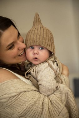 A woman holding a baby. High quality photo, The girl holds her child in her arms. The child is wearing a beige hand-knitted hat. mother and child are happy, they are smiling clipart