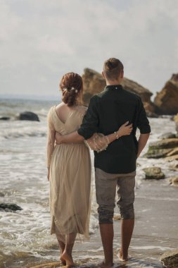 Deniz kıyısında yürüyen genç, sevgi dolu ve romantik bir çift. Arkadan bakıyorlar, birbirlerinin beline sarılıyorlar. ve sahilde yalınayak yürürken, kayalar önünde..