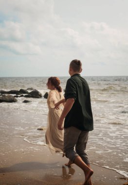 Sahilde koşuşturan aşık romantik bir çift. El ele tutuşuyorlar. Kız narin bir bej elbise giyiyor. Etraflarında deniz ve kayalar var. Hava çok açık. Arkadan görünüm