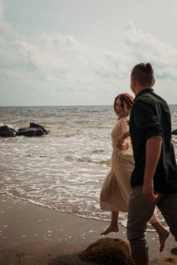 Sahilde koşuşturan aşık romantik bir çift. El ele tutuşuyorlar. Kız bej renkli bir elbise giyip adama bakıyor. Etraflarında deniz ve kayalar var. Hava açık.