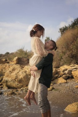 Romantik ve gülümseyen bir çift kızı kollarında tutar ve kaldırır. Çok duygusal bir fotoğraf. Yeni evliler mutlu ve gülümsüyor. Kız boynuna sarılıyor, o da onu kaldırıyor. Çevrelerinde de deniz ve kayalar.