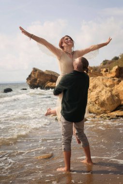 Romantik ve gülümseyen bir çift kızı kollarında tutar ve kaldırır. Çok duygusal bir fotoğraf. Yeni evliler mutlu ve gülümsüyor. Kız ellerini güneşe uzatıyor.
