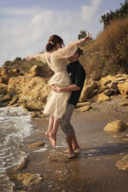 Romantik ve gülümseyen bir çift kızı kollarında tutar ve kaldırır. Çok duygusal bir fotoğraf. Yeni evliler mutlu ve gülümsüyor. Kız kollarını açtı, adam onu döndürdü. Çevrelerinde de deniz ve kayalar.