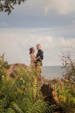 Güzel aşıklar bir uçurumun kenarında, gökyüzünün ve denizin zeminine karşı dururlar. Güneşli bir günde çekilmiş parlak bir fotoğraf. Kız saçını güzel bir topuz şeklinde yaptırıyor. Gülümsüyor.