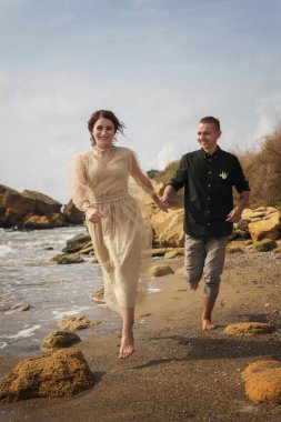 Aşıklar el ele tutuşup deniz kıyısında koşuyorlar. İleride bej elbiseli güzel bir kız var. Yeni evliler deniz ve kayaların arkasında kumların üzerinde yalınayak koşuyorlar. Açık renk pantolon giyen adam.