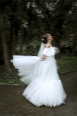 Beyaz elbiseli ve uzun peçeli bir gelin ağaçların arasında duruyor. Fotoğrafta gelin figürü bulanık. Dönüyor, dans ediyor ve gülüyor. Elbisesi ve duvağı dalgalanıyor.