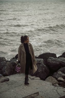 Kayaların etrafındaki deniz kıyısında genç bir kız. Bej renkli bir trençkot giymiş, deniz kıyısında kasımpatı çiçekleri arasında rüya görüyor. Mavi kumaşla süslenmiş bir sandalye.