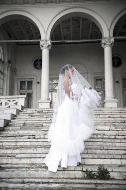 Bir buket çiçekle kaplanmış güzel bir gelin. Güzel bir kız, aydınlık bir binanın merdivenlerinde duruyor. Beyaz, uzun bir elbiseyle yan yana duruyor. 