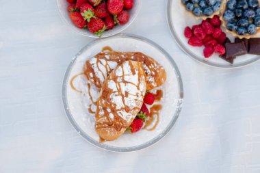 Ortaya taze çilek serpiştirilmiş mahar tozu serpiştirilmiş iki kruvasan. Yanlarındaki tabakta taze çilekler ve taretlerde çikolata parçalı yabanmersinleri var.
