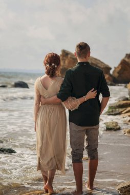 Deniz kıyısında yürüyen genç, sevgi dolu ve romantik bir çift. Arkadan bakıyorlar, birbirlerinin beline sarılıyorlar. ve sahilde yalınayak yürürken, kayalar önünde..