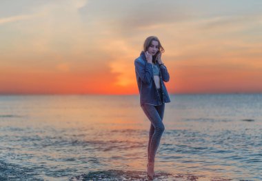 a slender girl stands on the beach of the sea at sunrise. she is dressed in tight jeans and stands at full height. the sea is calm without waves. she stands on a stone clipart