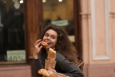 Sokakta ekmek yiyen güzel genç bir kadın. Bir kafede elinde baget ekmeğiyle oturuyor. Gülümsüyor. Kabarık saçları var.