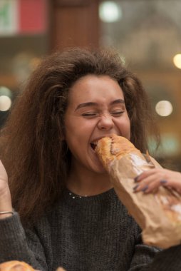 Genç bir kız iştahla baget yer. Dalgalı kabarık saçları var. Ekmek masada yatıyor. Yüze yakın çekim. Tarafsız gri kıyafetler giyiyor ve parlak duygular gösteriyor.