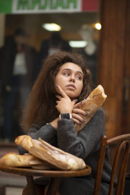 Genç bir kız bir kafede bir masada oturuyor ve bir ekmeği var. Kız başka tarafa bakıyor. O, arkadaşını bekliyor. Üç parça baget ekmeği var. Öğrencinin dalgalı kabarık saçları var..