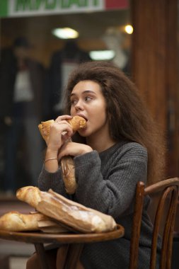 Genç bir kız bir kafede bir masada oturuyor ve bir baget yiyor. Bir baget yiyor ve onu ısırıyor. Onun önündeki masada üç baget ekmeği var. Öğrencinin dalgalı kabarık saçları var..