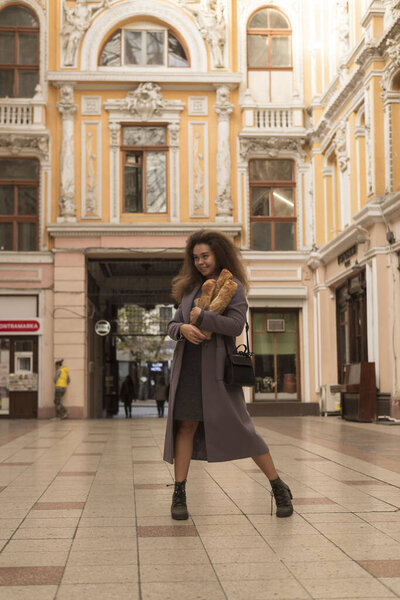 Красивая девушка в пальто ходит по городу, держа в руках буханку хлеба. Девушка держит французский рулет. У неё пушистые вьющиеся волосы и тёмно-серое пальто, ходит по игровым автоматам