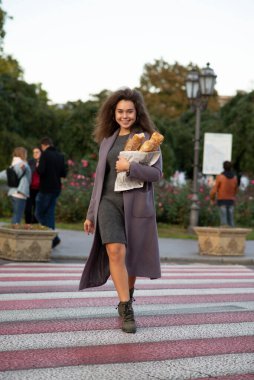 Kız elinde üç baget tutuyor. Şehirde yürüyor, kameraya bakıyor ve güzel bir gülümsemesi var. O bir ceket ve bot giyiyor. Yaya geçidinden karşıya geçiyor..