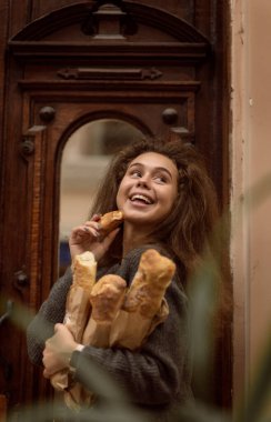 Kızın baget ekmeği var. Ortalama çekim, kameraya gülümsüyor. Ellerinde üç baget ve bir tane var.